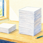 机の上に高さの違う書類の山が2つ。右側の山が明らかに高く積み上がり、そばにコーヒーカップとペンが置かれている