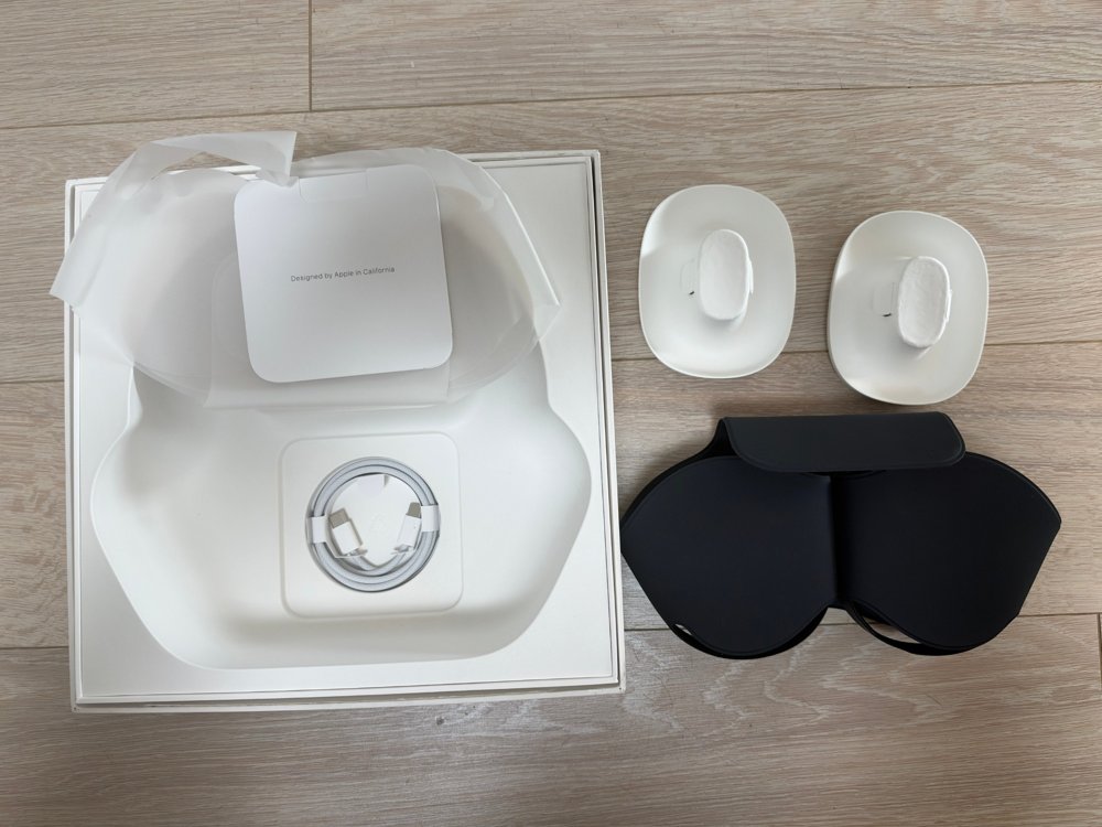 AirPods Maxの外箱の中敷き、白い編み込みの充電ケーブル、説明書類の袋、白い中敷き2つ、黒いSmart Caseを床に並べた写真